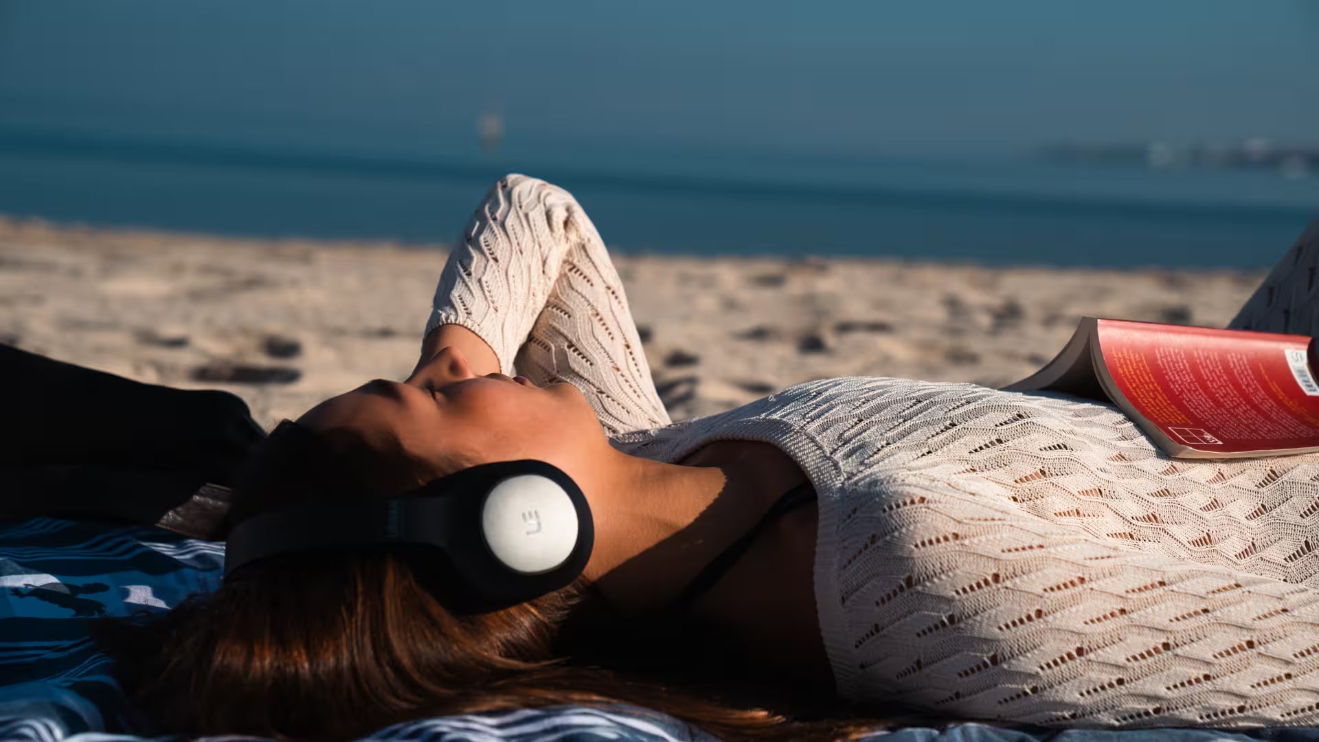A woman lying on a beach blanket on a sandy beach, wearing white over-ear headphones and a white knit top, with a red book in her hand. The background shows the blue ocean and sky.