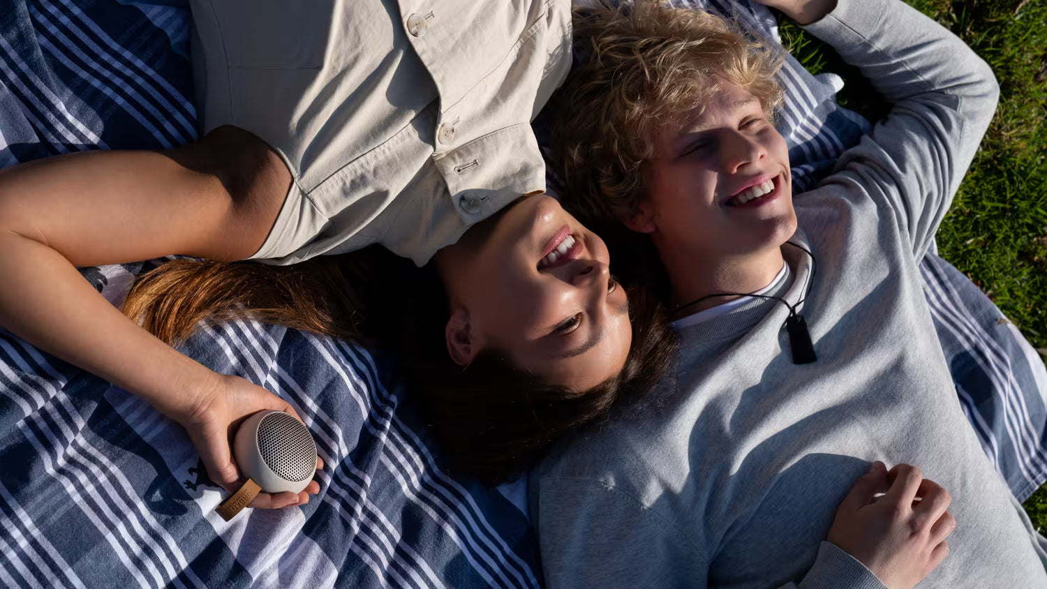 Two young adults lying on a blue and white striped blanket on grass, smiling and talking. One holds a small round portable speaker.