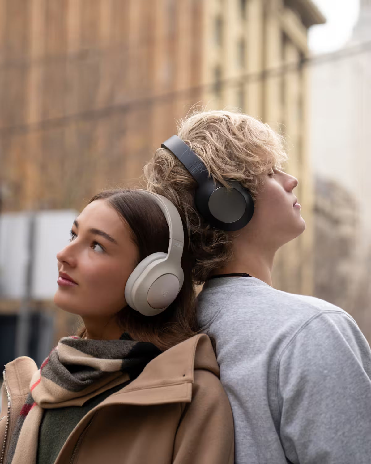 Two people standing back-to-back outdoors, each wearing over-ear headphones. The person on the left has light brown hair and wears a beige coat and striped scarf, while the person on the right has wavy blond hair and wears a gray sweatshirt.