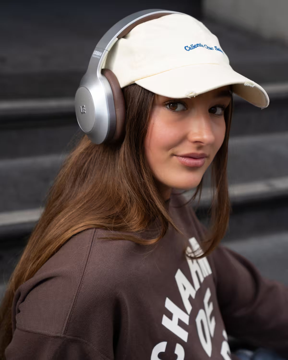 A young woman with long brown hair is shown in a lifestyle setting, wearing over-ear headphones, a cream baseball cap with blue text, and a brown sweatshirt. The background consists of dark gray concrete steps.