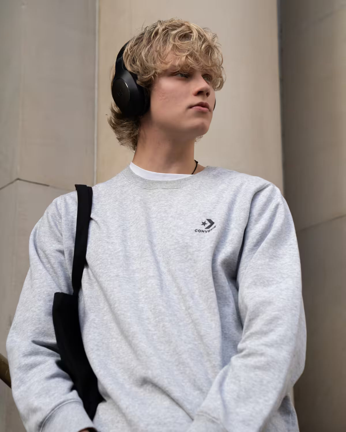 A young man with blonde hair wearing a light gray heather crewneck sweatshirt with a black Converse logo on the chest, black over-ear headphones, and a black shoulder bag, standing against a neutral beige stone wall.