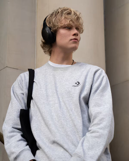 A young man with blonde hair wearing a light gray heather crewneck sweatshirt with a black Converse logo on the chest, black over-ear headphones, and a black shoulder bag, standing against a neutral beige stone wall.