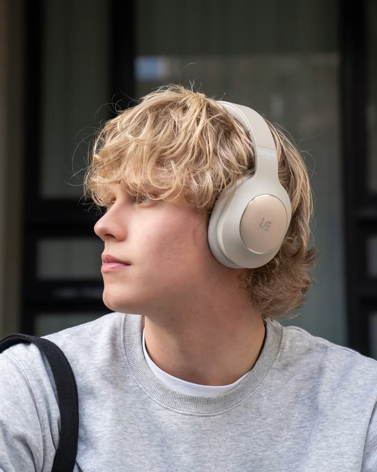 A young man with blonde hair wearing light beige over-ear headphones and a gray sweatshirt, shown in profile against a dark windowed background.