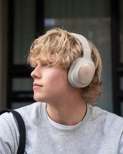 A young man with blonde hair wearing light beige over-ear headphones and a gray sweatshirt, shown in profile against a dark windowed background.